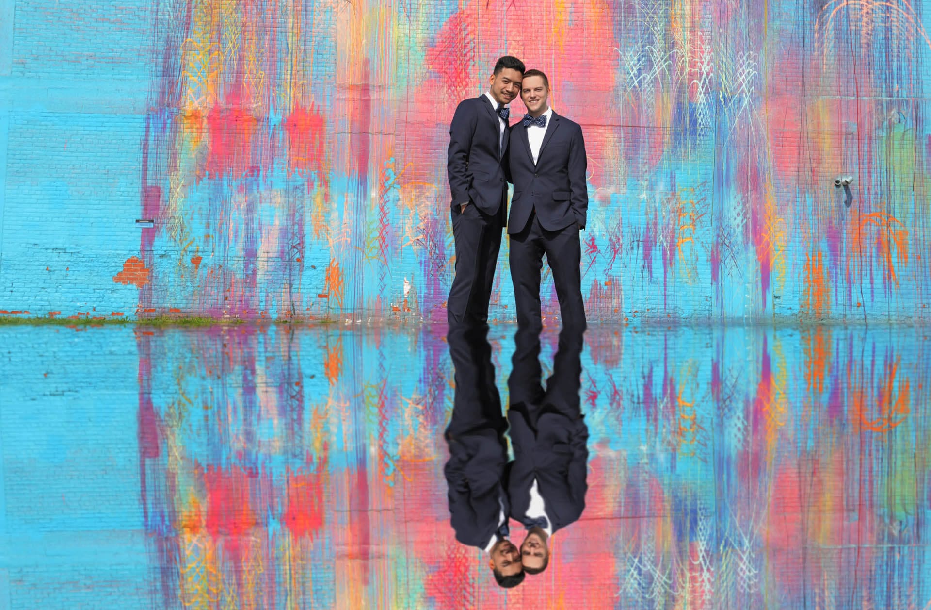 Two grooms are reflected in a puddle with the bleeding rainbow wall in Detroit behind them after their same sex gay wedding.