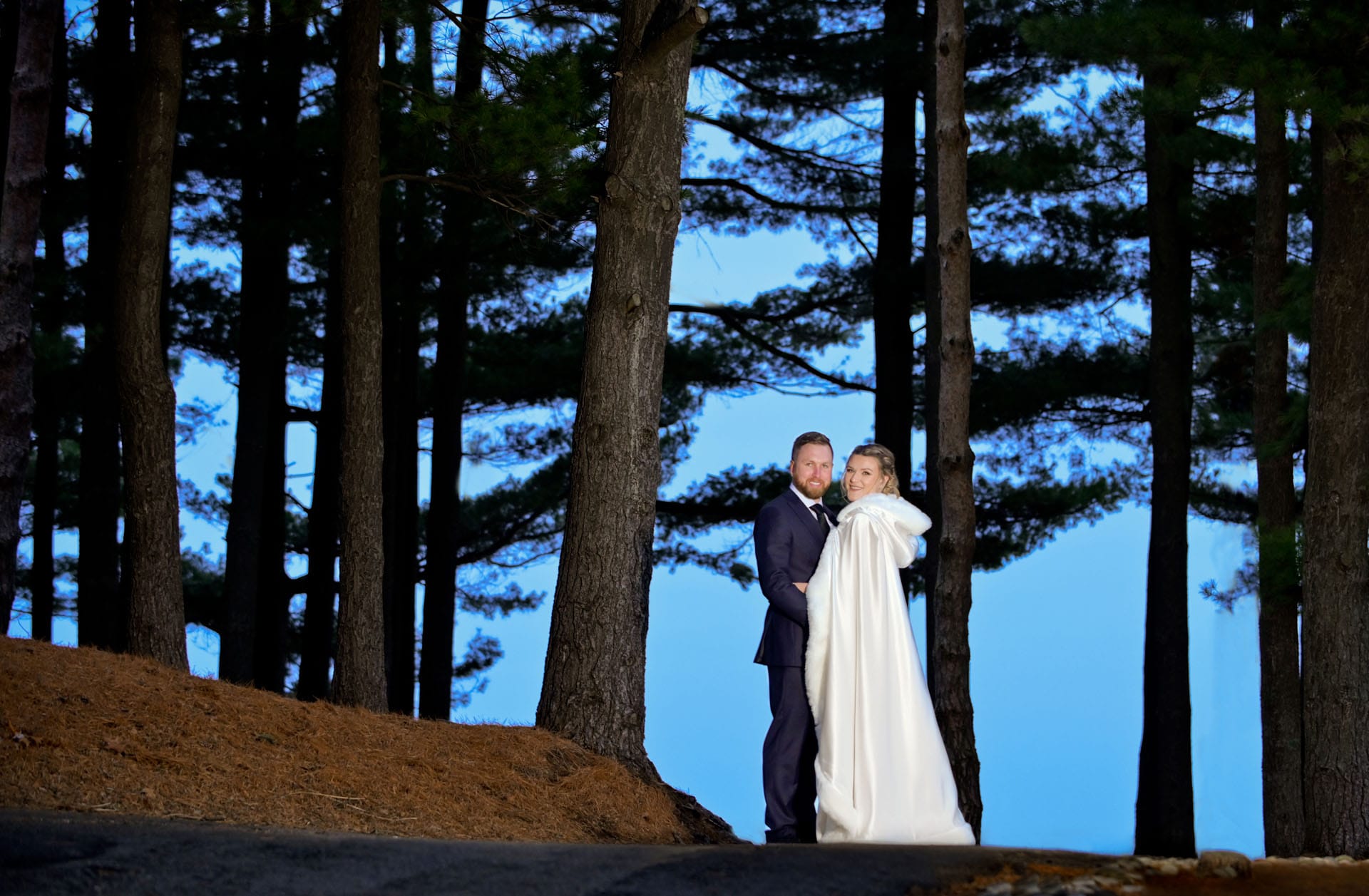 Bride and groom pose in woods at Oakhurst Golf Course in Clarkston, Michigan in winter at dusk.