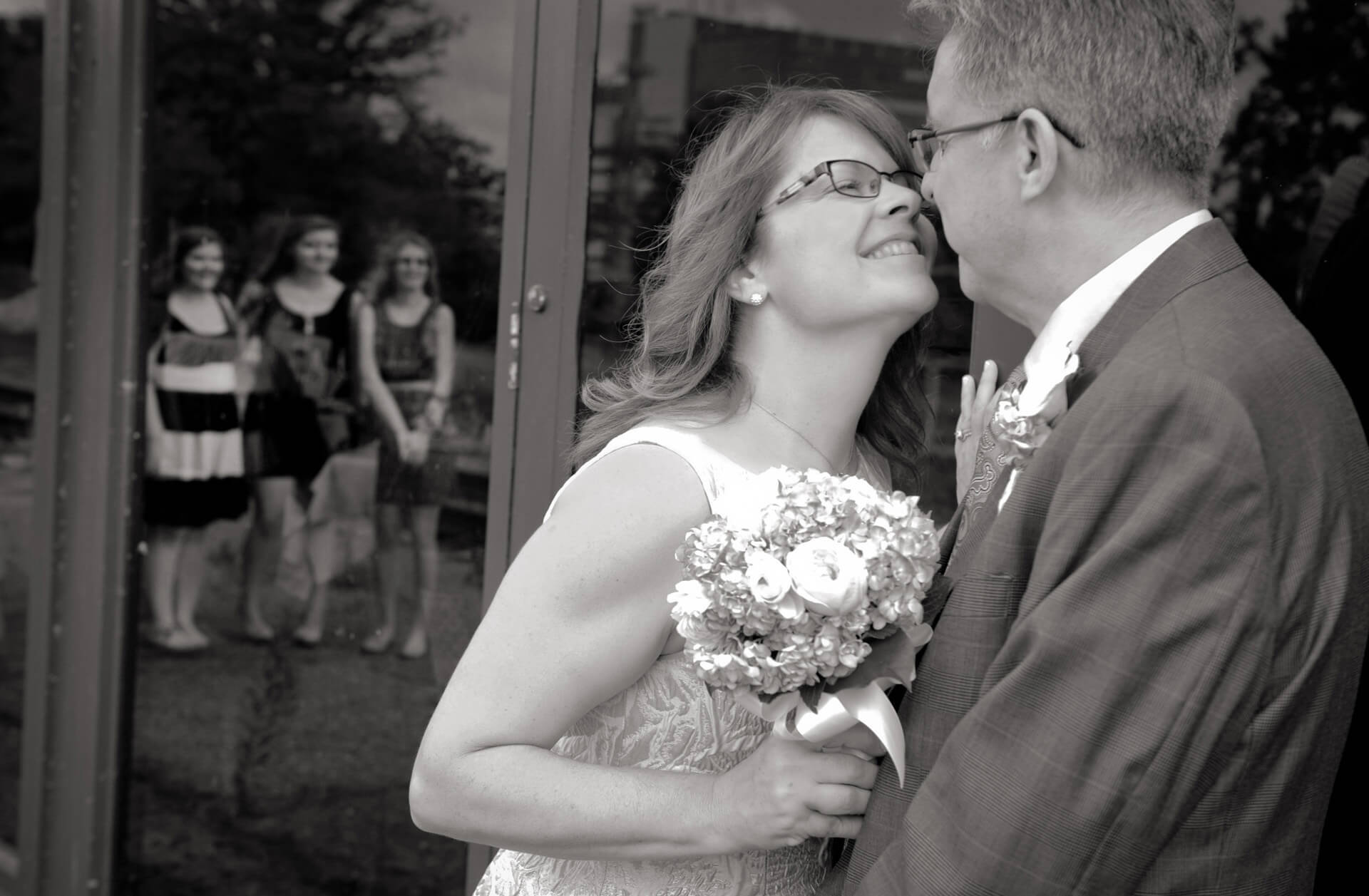The bride and groom kiss while their kids look on in the reflection of the depot at the Gandy Dancer in Ann Arbor, Michigan.